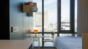 Une chambre d’hôtel comprenant un lit, une petite table avec une chaise, une fenêtre donnant sur des immeubles et une étagère avec divers objets.