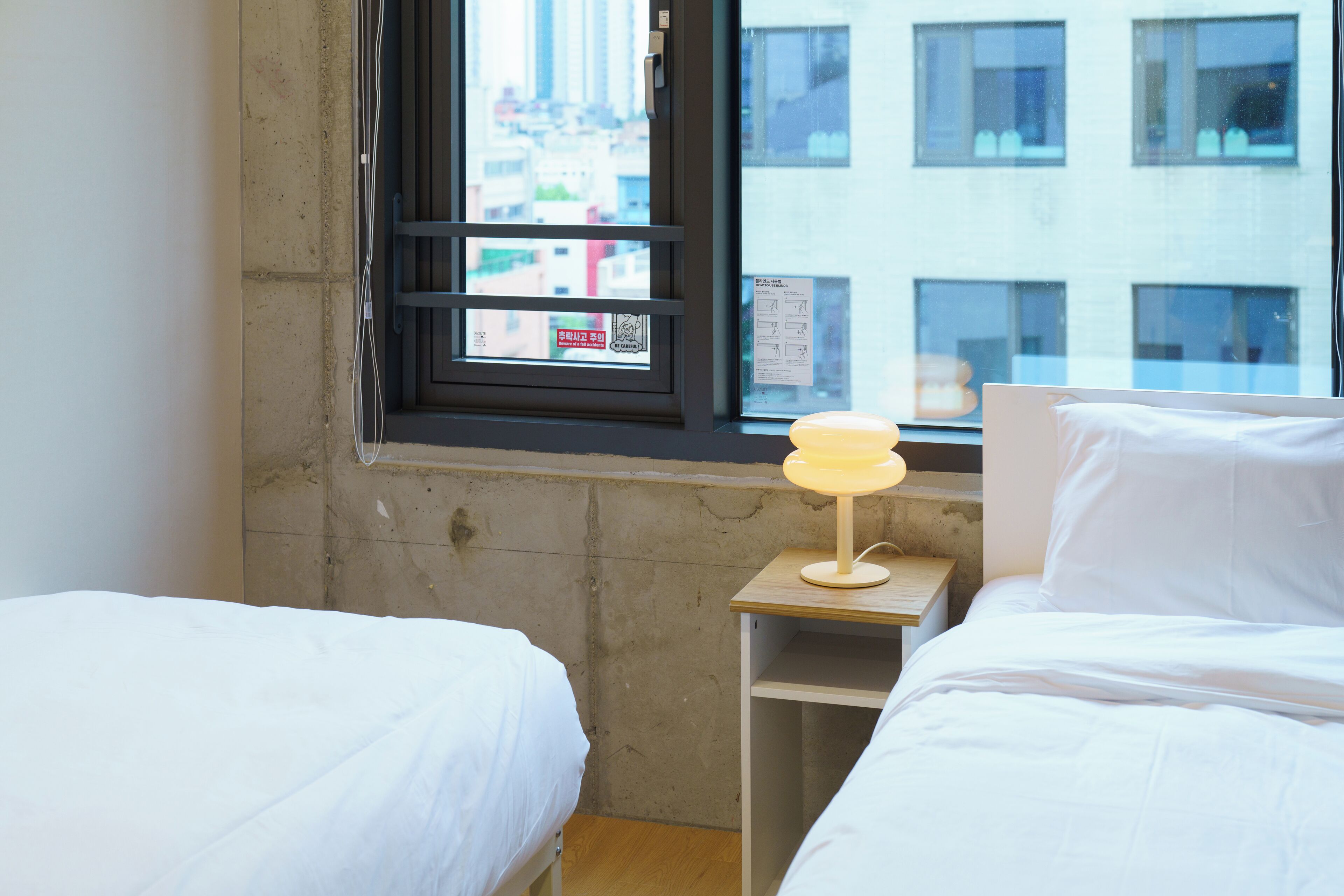 Habitación de hotel con dos camas, una ventana que ofrece vistas a edificios y una pequeña mesa con una lámpara.