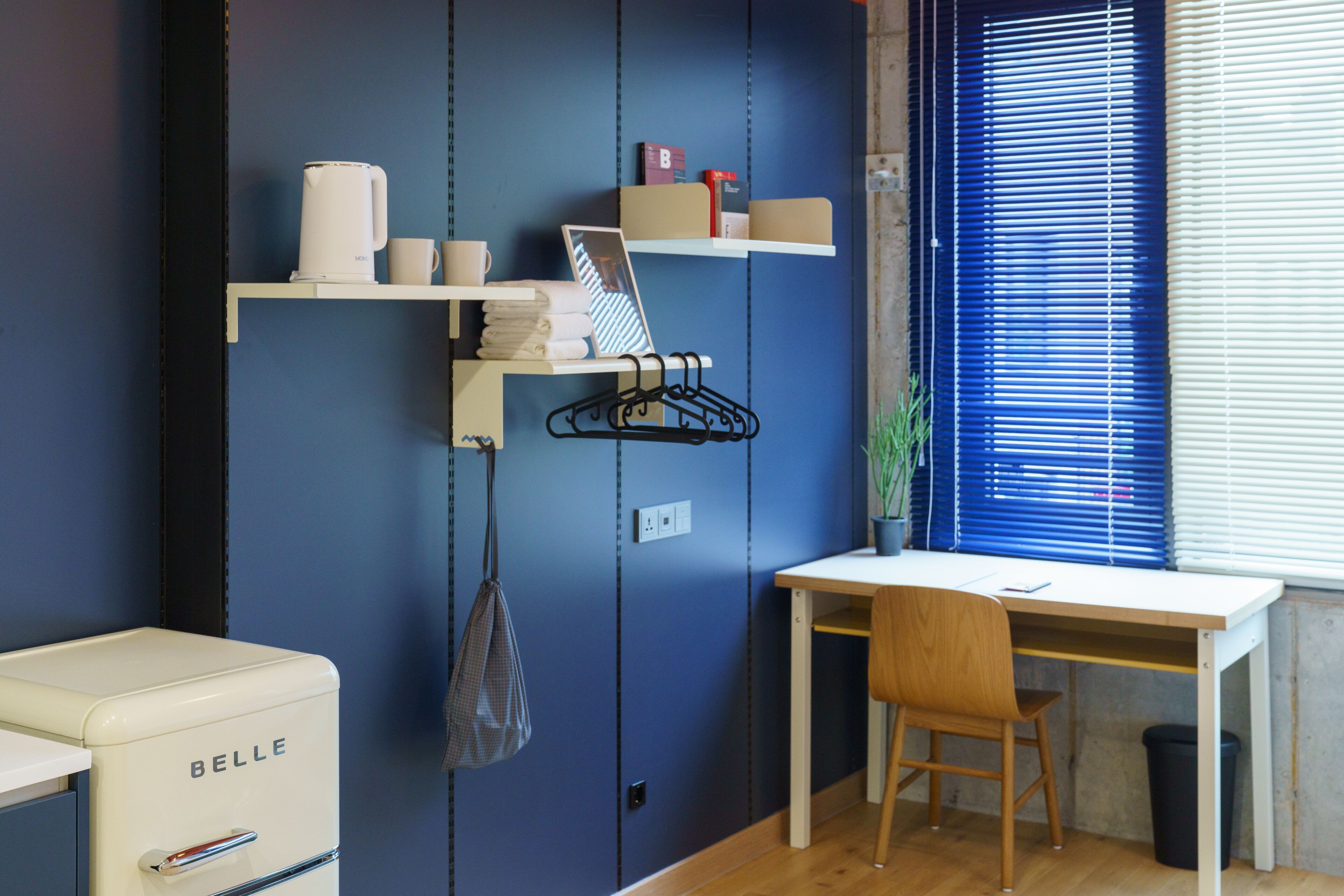 A room with a desk, chair, and a white appliance labeled 'BELLE'. There are shelves with items and a window with blinds.