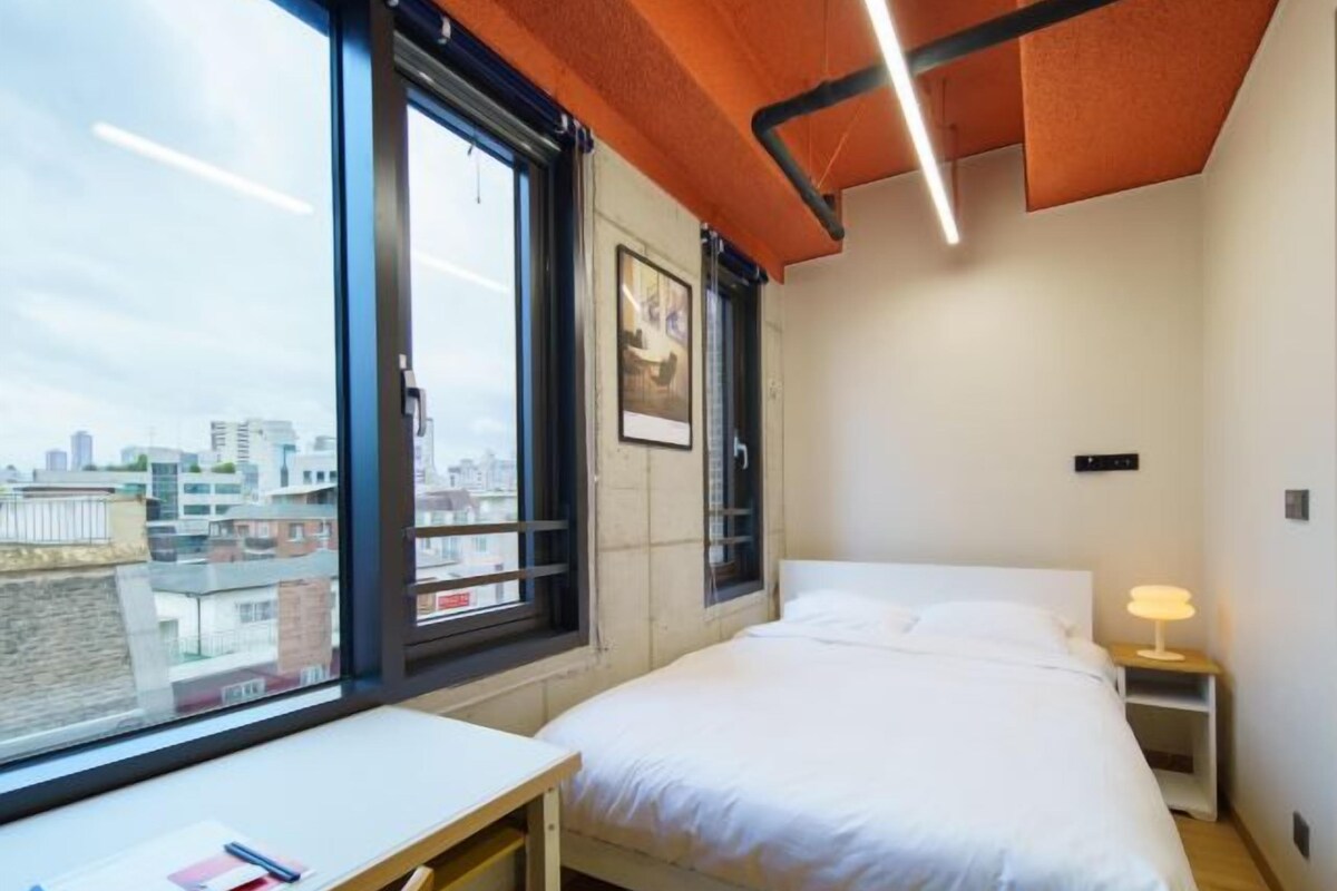 A modern bedroom with a bed, a desk, and a window with a city view.