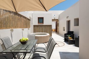 Un patio al aire libre con mesa de vidrio, sillas, una zona de estar circular y una pequeña mesa con bebidas.