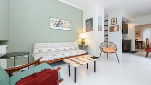 Ein modernes Wohnzimmer mit einer Couch, einem Couchtisch und einem Sessel. Eine Lampe steht auf einem Beistelltisch, und im Hintergrund ist eine Küchenzeile zu sehen.