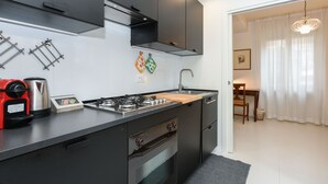 Eine moderne Küche mit Gaskochfeld, Backofen und Kaffeemaschine. Durch eine offene Tür ist eine Essecke mit Tisch und Stühlen zu sehen.