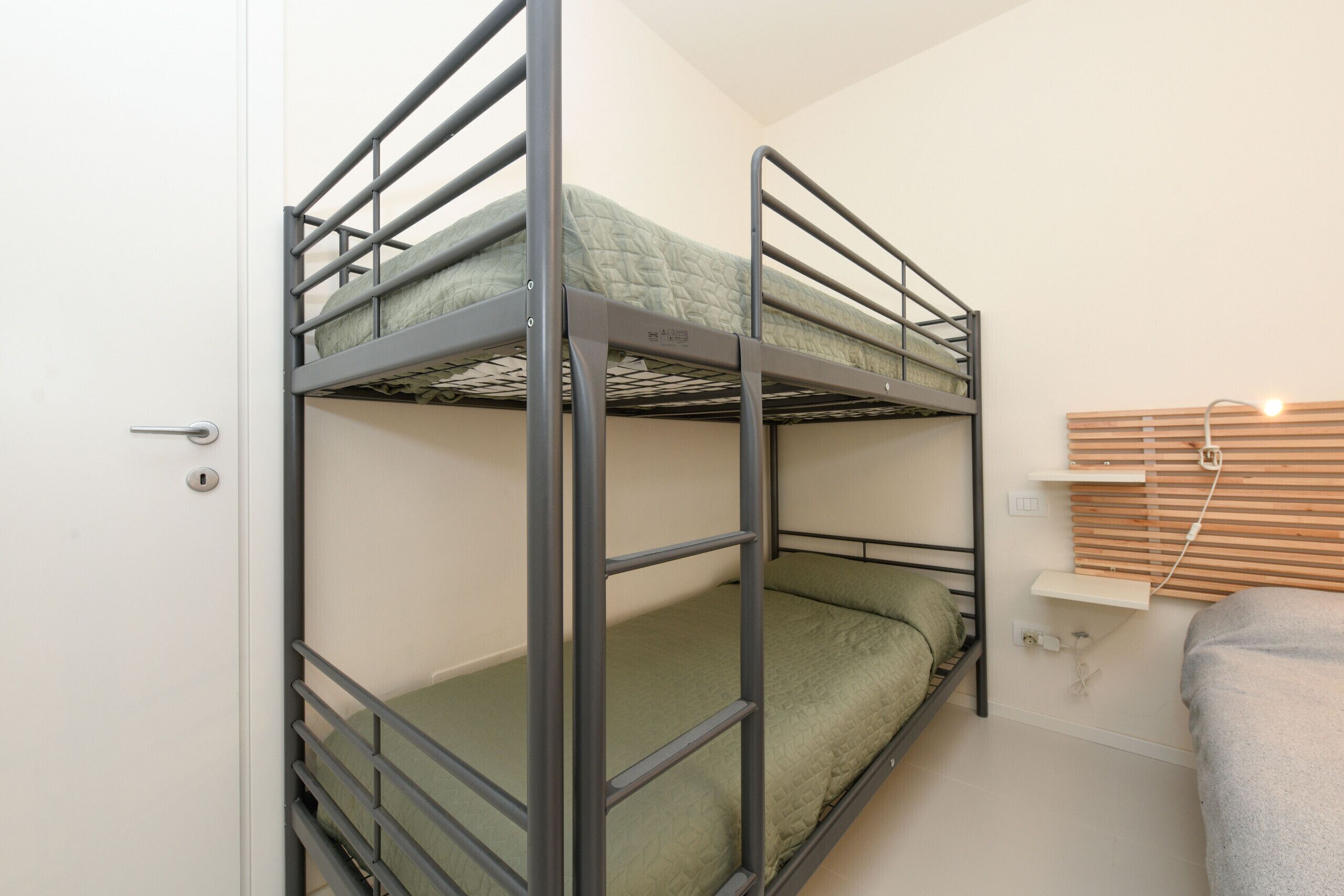Ein Stockbett mit Metallgestell und grüner Bettwäsche, ein Holzregal mit Steckdose und eine Tür mit Schloss.