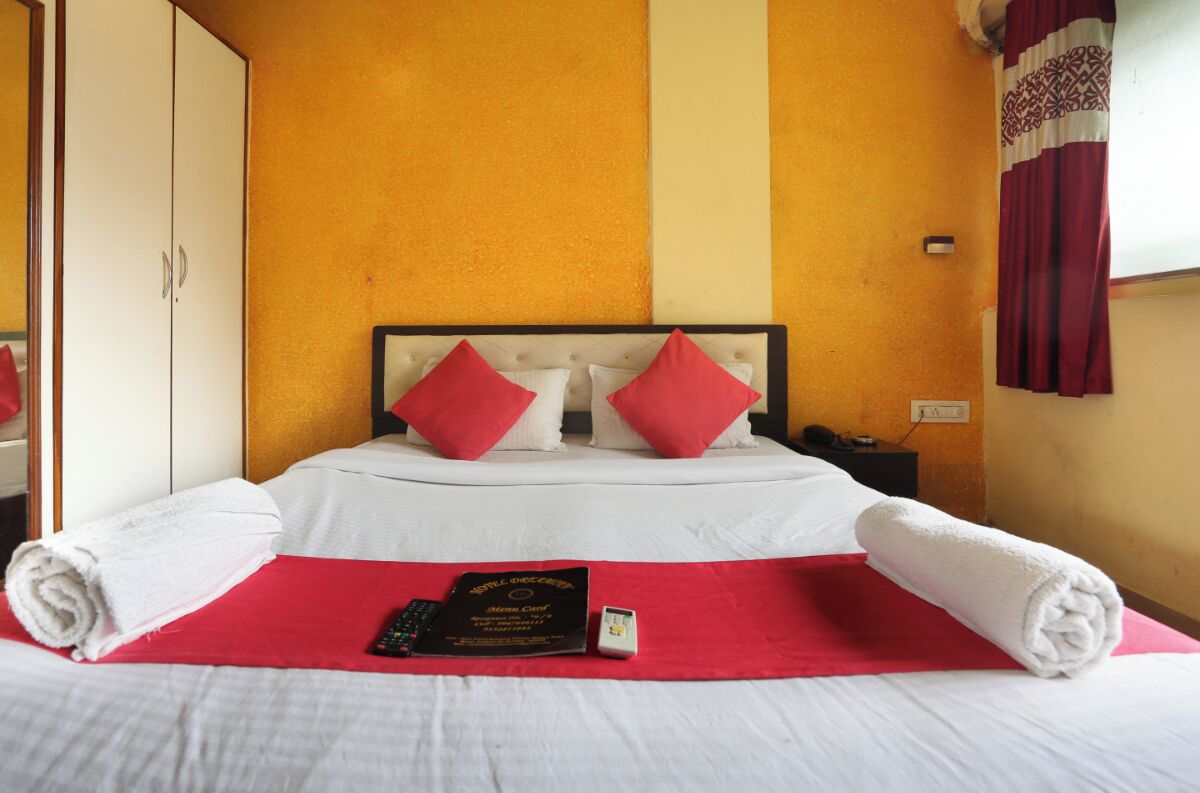 Une chambre d’hôtel avec un lit, des oreillers, une couverture rouge, une serviette, une télécommande, un livre et une prise électrique au mur.