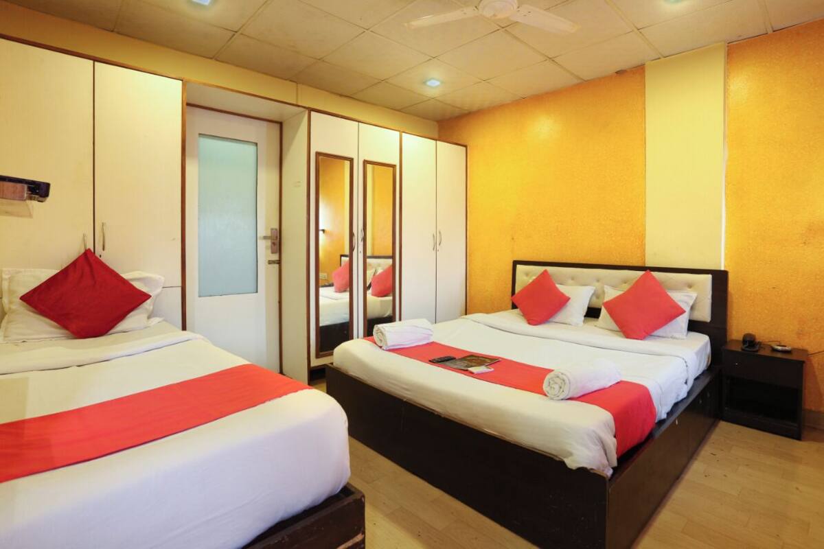 Une chambre d’hôtel avec deux lits, chacun agrémenté d’oreillers rouges et d’une couverture rouge. On y trouve une armoire avec un miroir, une table de chevet avec une lampe et une petite table de nuit avec un téléphone.