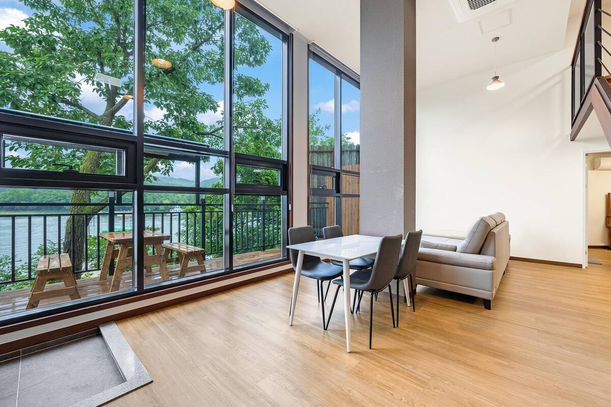 Amplia habitación con grandes ventanales que ofrecen vistas a un lago, una mesa de comedor de madera con sillas, un sofá y una escalera.