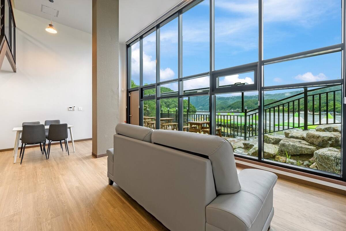 Un interior moderno con un sofá gris, suelo de madera, zona de comedor con mesas y sillas, y ventanas amplias con vistas a un paisaje montañoso.
