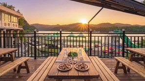 Une terrasse en bois avec une table dressée pour le repas, donnant sur une étendue d’eau et un port de plaisance où sont amarrés des bateaux.