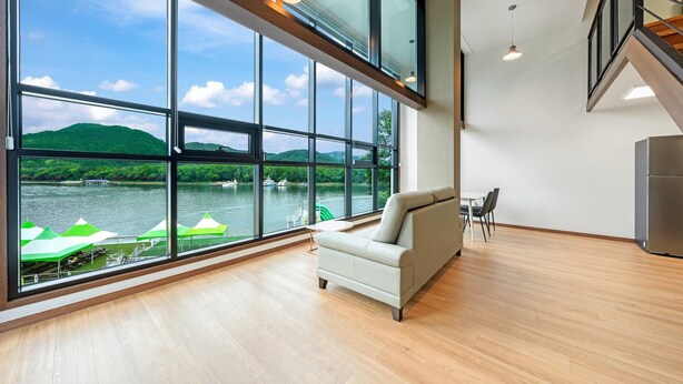 Una habitación moderna con amplios ventanales con vistas a un lago, un sofá, una mesa de comedor y un refrigerador.
