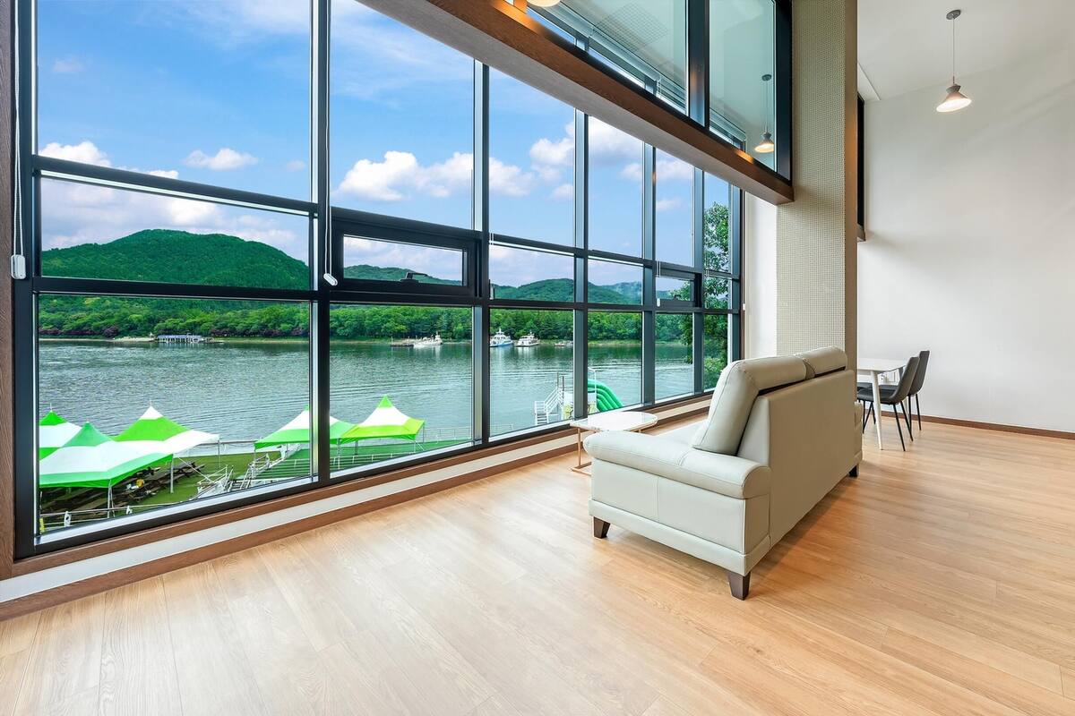 Una habitación moderna con amplios ventanales con vistas a un lago, un sofá, una mesa de comedor y un refrigerador.