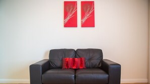 Un sofá de cuero negro con un cojín rojo en una habitación con piso de madera y dos obras de arte abstracto rojas en las paredes.