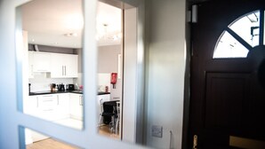 Vista a través de una puerta que da a una cocina moderna con armarios blancos, una zona de comedor con sillas y un ventanal sobre la puerta.