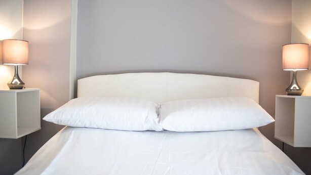Una cama bien tendida con sábanas y almohadas blancas, flanqueada por dos mesitas de noche con lámparas.