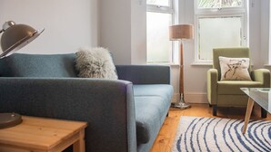 A living room with a blue sofa, a green armchair, a wooden side table, a floor lamp, and a patterned rug. - Rugby Lodge 3 Bedrooms Dublin (Dublin)