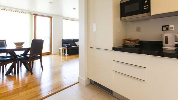 A modern kitchen with white cabinets, a black countertop, and a microwave. There is a dining area with a table and chairs, and a sofa in the background. - Grande Central Sandyford (Dublin)