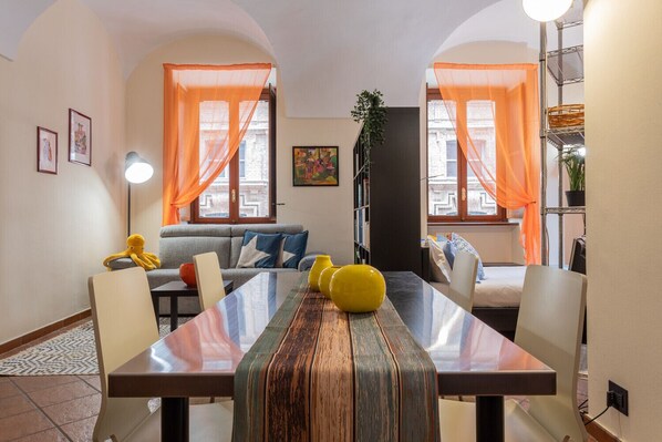 A dining area with a long table, chairs, and a sofa. There are two windows with orange curtains, a bookshelf, and a lamp.