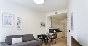 Un appartement moderne avec un canapé gris, une table à manger avec des chaises, un coin cuisine et des œuvres d’art encadrées accrochées aux murs.