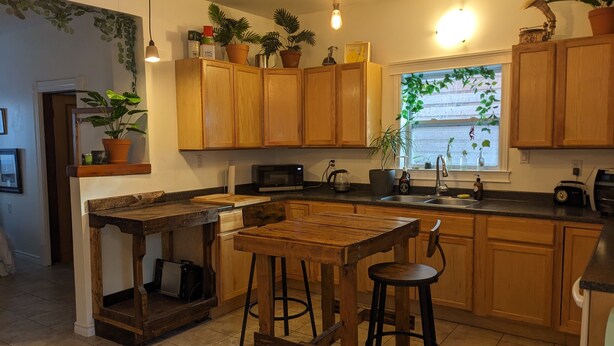 Una cocina con armarios de madera, una isla central con taburetes, un microondas y varias plantas en macetas.