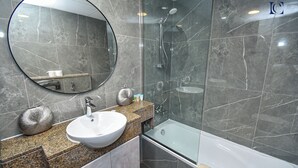 A modern bathroom with a granite countertop, a white sink, a round mirror, a glass-enclosed shower, and a bathtub. - Brand New Studio with Marina View -PAU (Dubai)