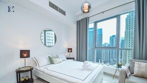 A modern hotel room with a large bed, a round mirror, a city view, and a small side table with decorative items. - Brand New Studio with Marina View -PAU (Dubai)