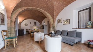 A spacious living area with a kitchen in the background, featuring a brick archway, a grey sofa, a white armchair, and a dining table with chairs. - Villa Le Ripe (Gambassi Terme)