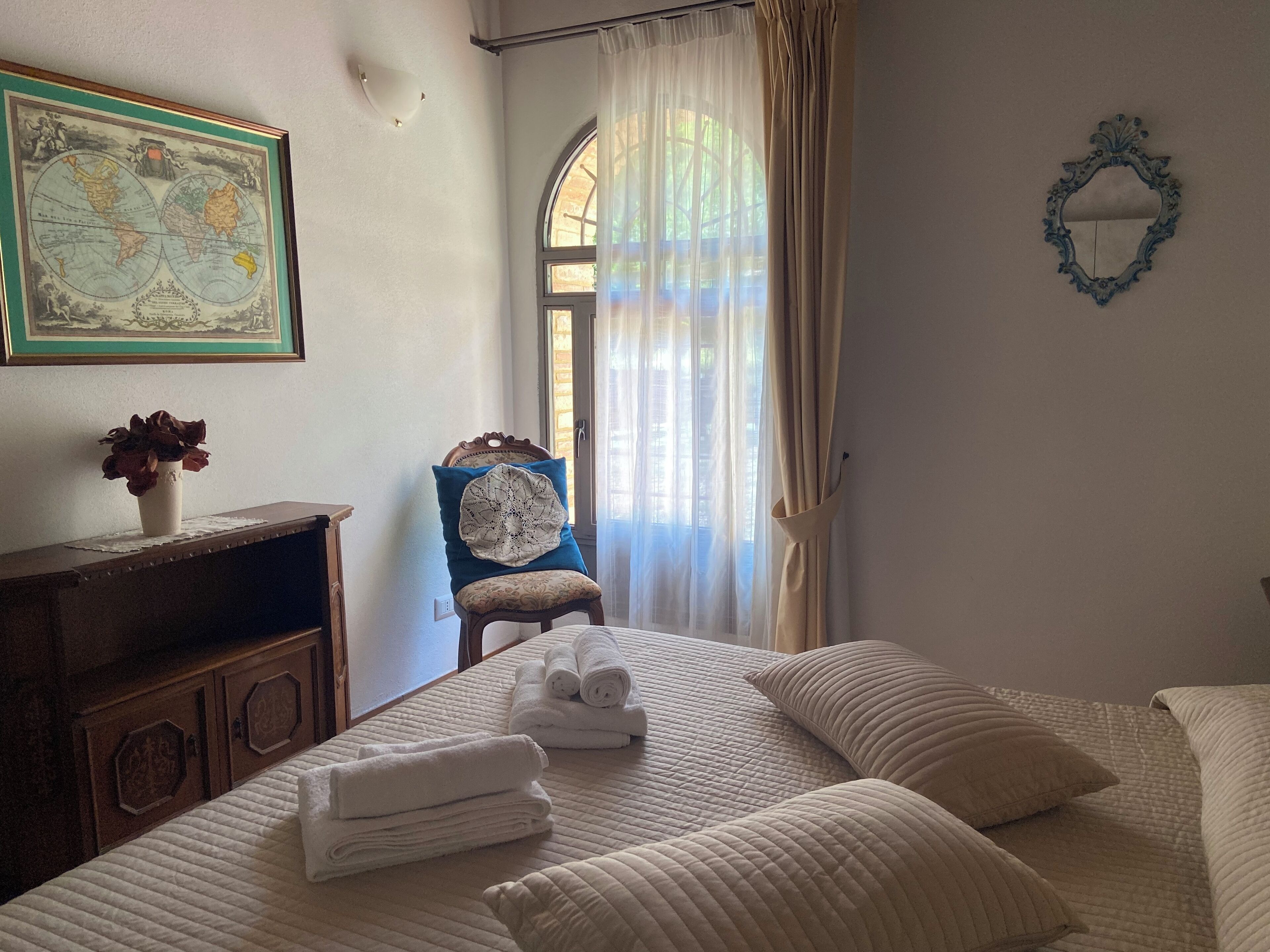 A neatly arranged bedroom with a bed, pillows, a chair, a map on the wall, and a window with curtains.