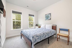 Une chambre à coucher comprenant un lit, deux fenêtres, une table de chevet, une chaise et une œuvre d’art accrochée au mur.