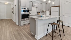 Una cocina moderna con armarios blancos, horno empotrado, una isla grande con taburetes e iluminación colgante.