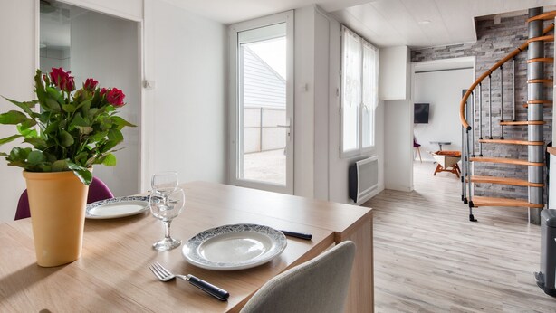 A dining area with a table set for two, a vase with flowers, and a view of the outside through a large window.