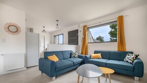 A modern living room with a blue sofa set, a round coffee table, and a view of the outdoors through a large window. - Tour-du Parc - Modern house for 8 (Le Tour-du-Parc)