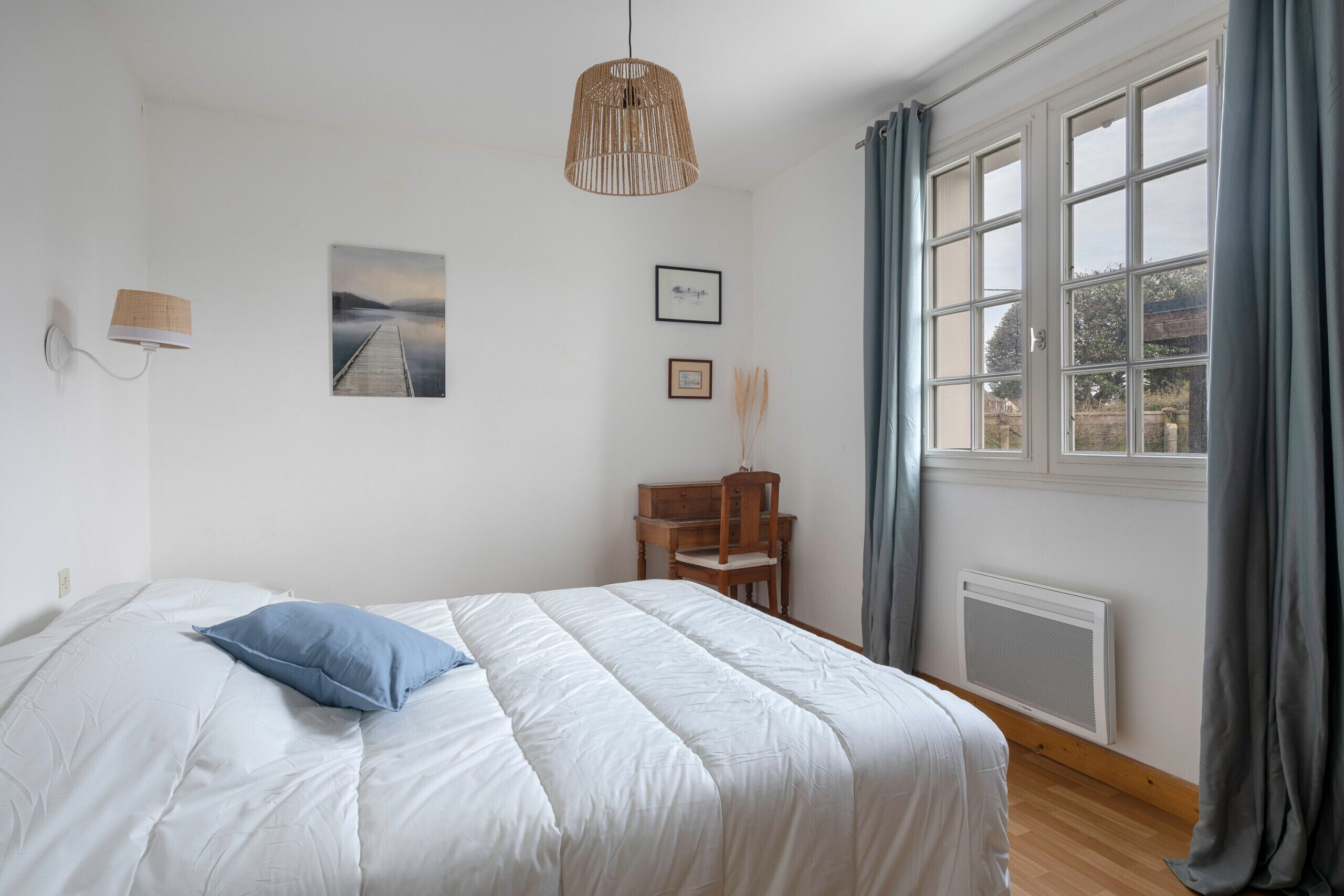 Un dormitorio con cama, escritorio, silla, ventana con cortinas, radiador y luces fijadas a la pared.