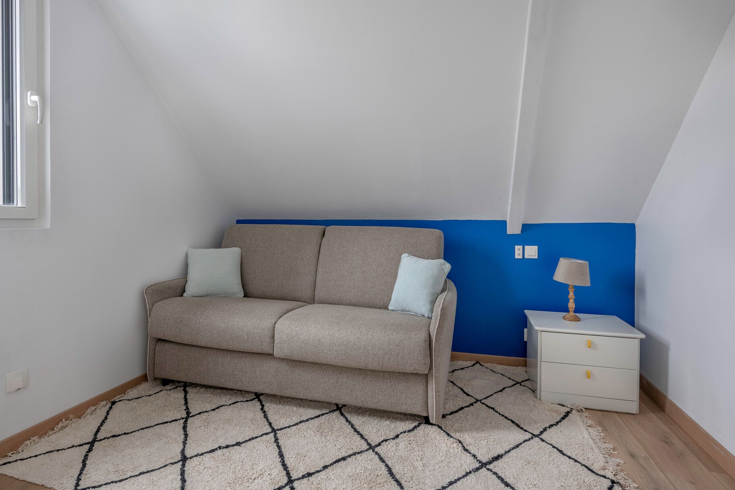 Een kleine, gezellige kamer met een beige bank, een wit nachtkastje en een blauwe accentmuur.