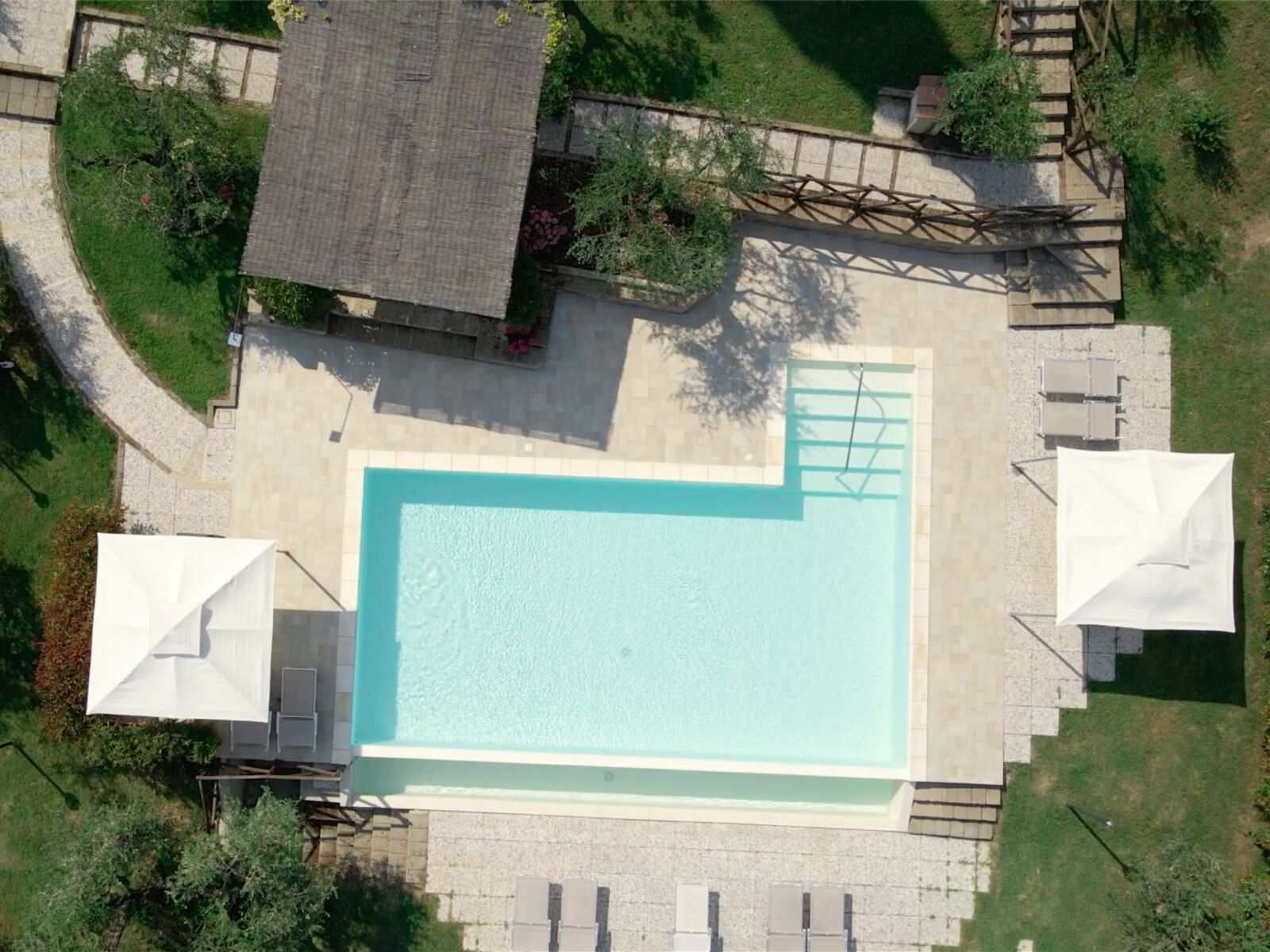 An aerial view of a rectangular swimming pool with a tiled deck surrounding it, a small covered seating area, and a wooden deck with steps leading to a raised platform.