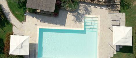 An aerial view of a rectangular swimming pool with a tiled deck surrounding it, a small covered seating area, and a wooden deck with steps leading to a raised platform.