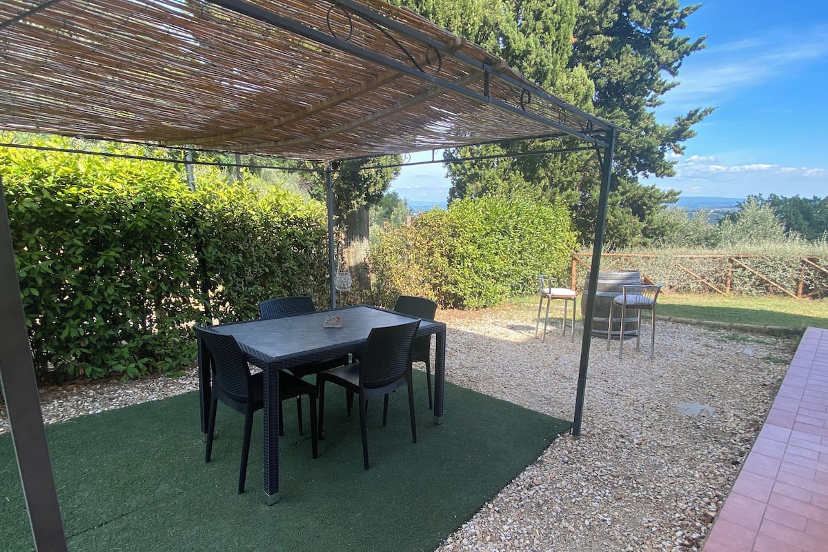 A covered patio with a table and chairs, surrounded by greenery and a view of the landscape.