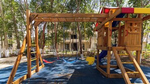 Une structure de jeux en bois, comprenant un toboggan, des balançoires et une zone d’escalade, entourée d’arbres et recouverte d’un sol bleu.