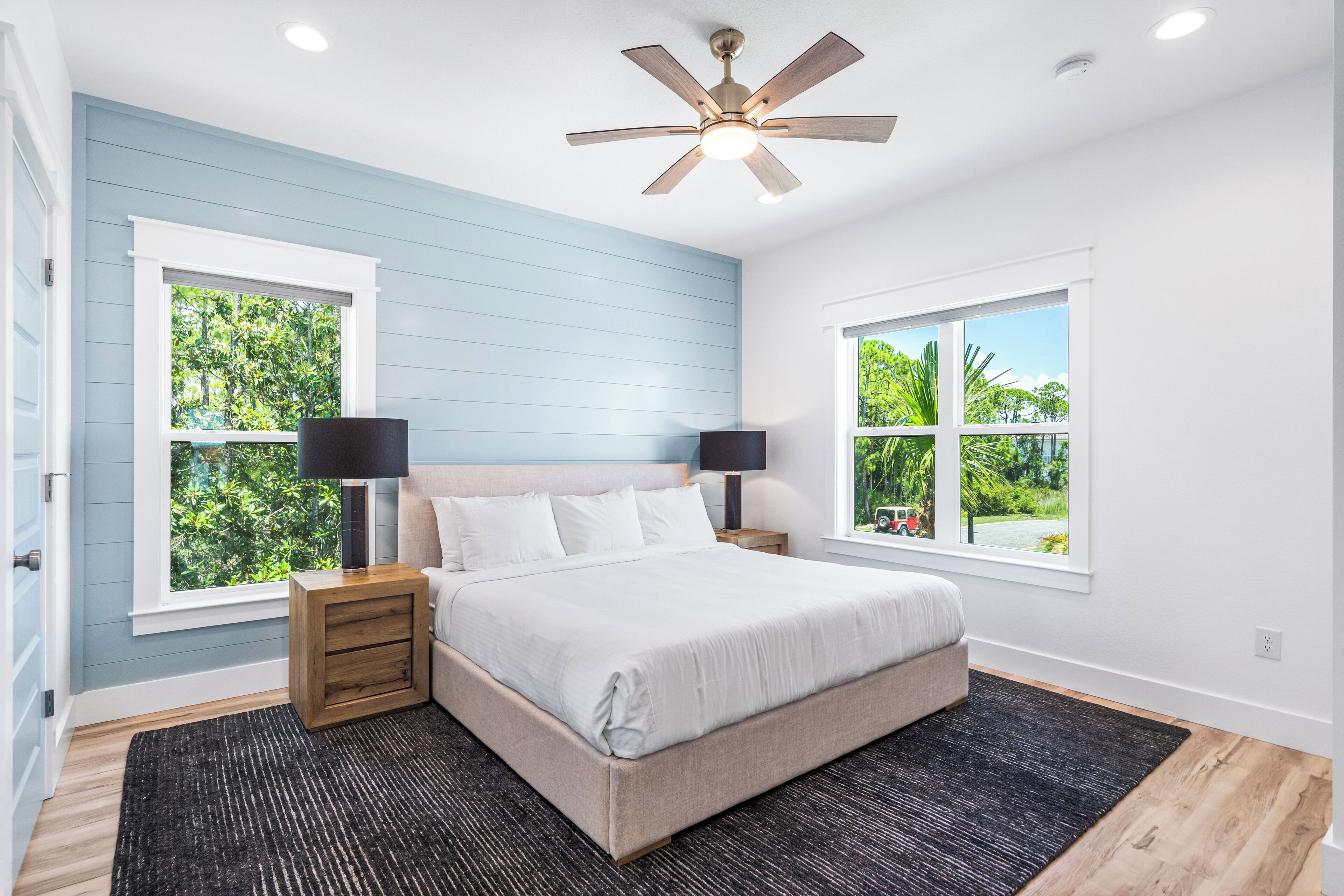 A bedroom with a bed, two bedside tables with lamps, a ceiling fan, and two windows with views of greenery.