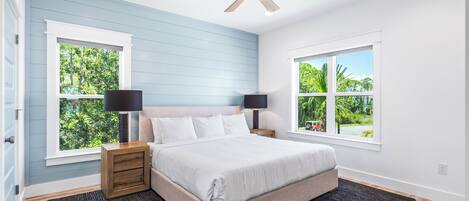 A bedroom with a bed, two bedside tables with lamps, a ceiling fan, and two windows with views of greenery.