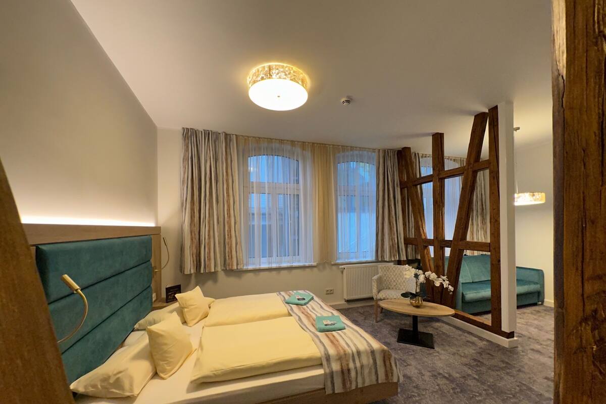 Habitación de hotel con cama, escalera de madera, mesita con un florero y ventana con cortinas.