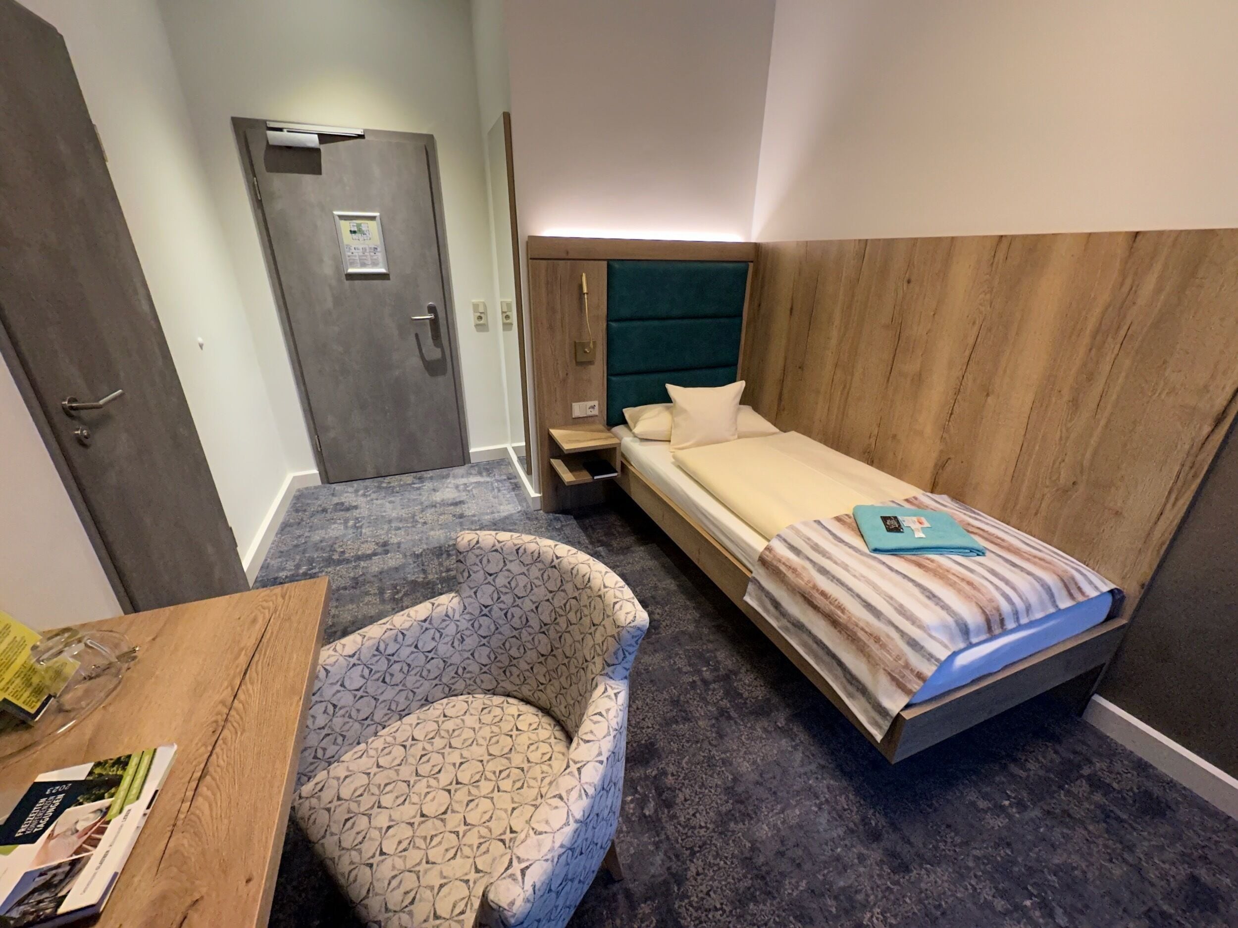 Una habitación de hotel con una cama, una silla, un escritorio y una revista, además de una puerta.