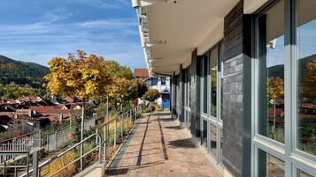 Uma varanda com caminho de tijolos, guarda-corpo de metal e grandes janelas com vista para uma área residencial com folhagem de outono.