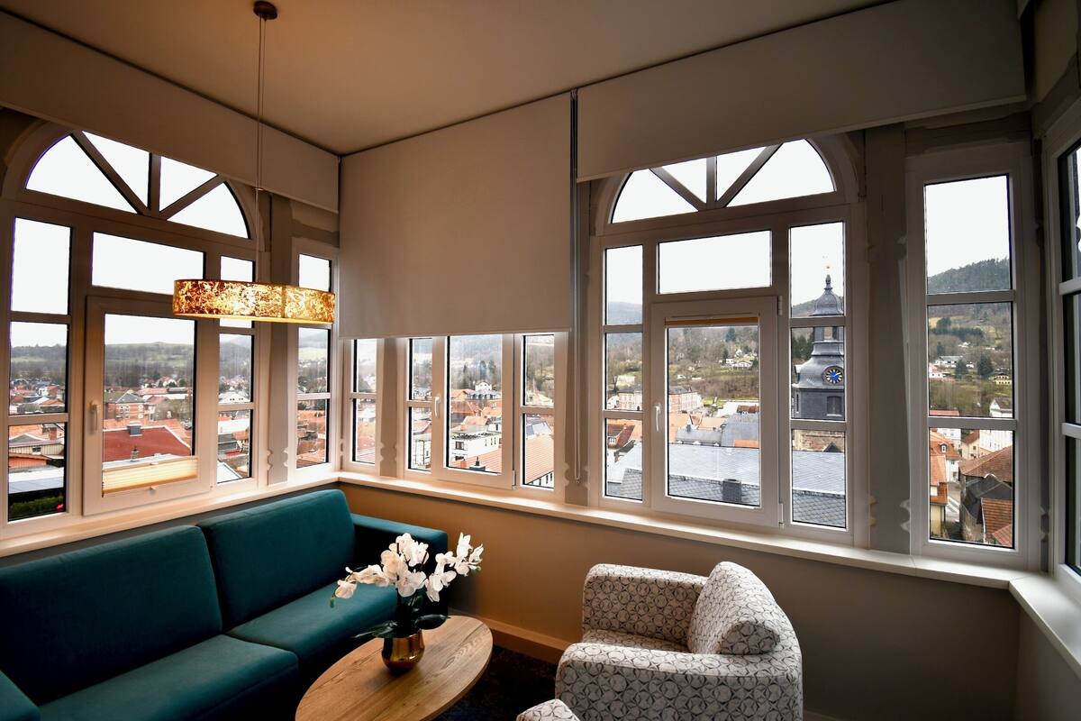 Una habitación con un sofá azul verdoso, un sillón estampado y una mesa de centro de madera con flores. Amplias ventanas ofrecen vistas a un pueblo y a la aguja de una iglesia.