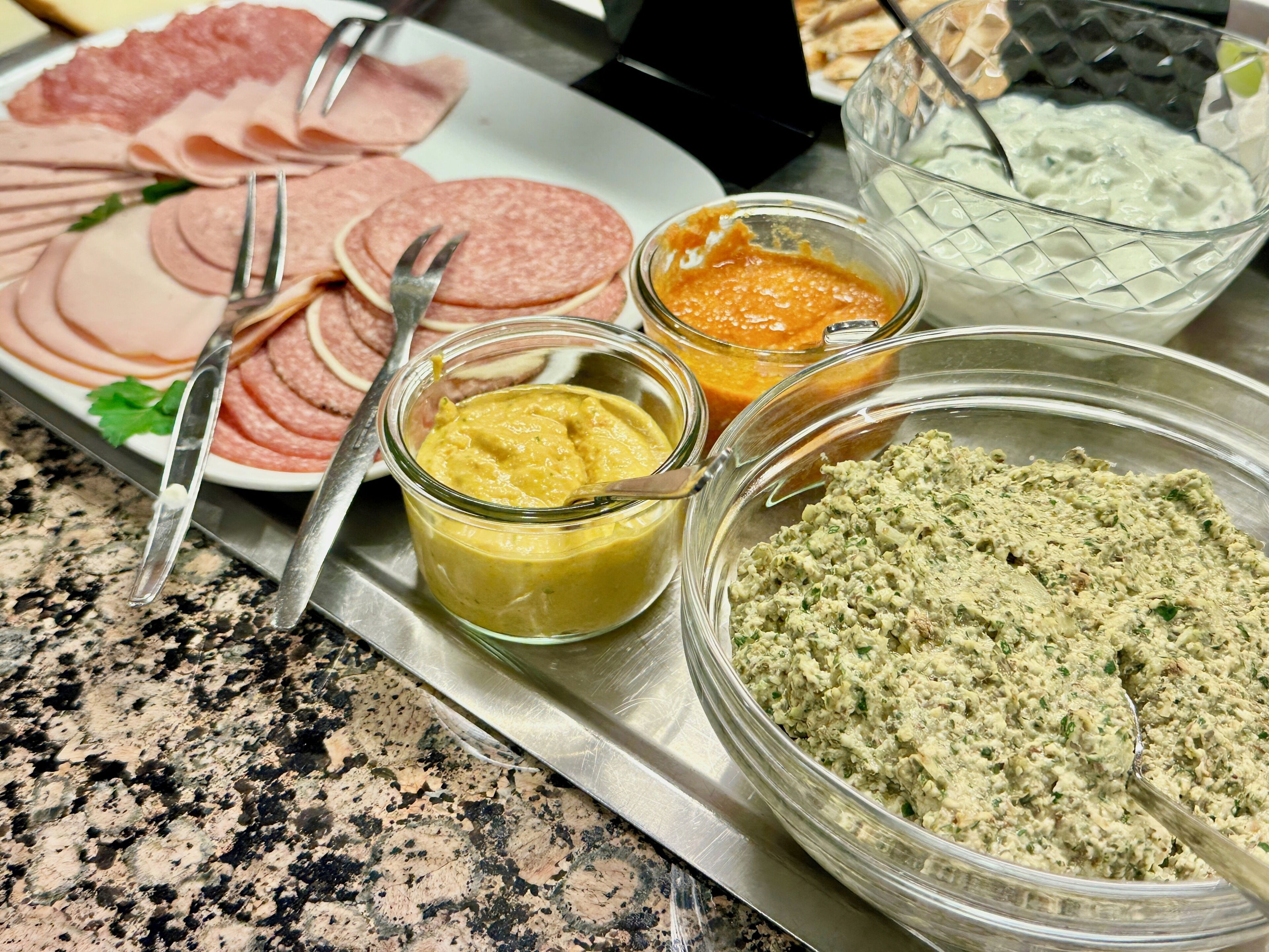 A buffet table with various food items including sliced meats, a bowl of green dip, a jar of yellow spread, and a container of orange sauce.