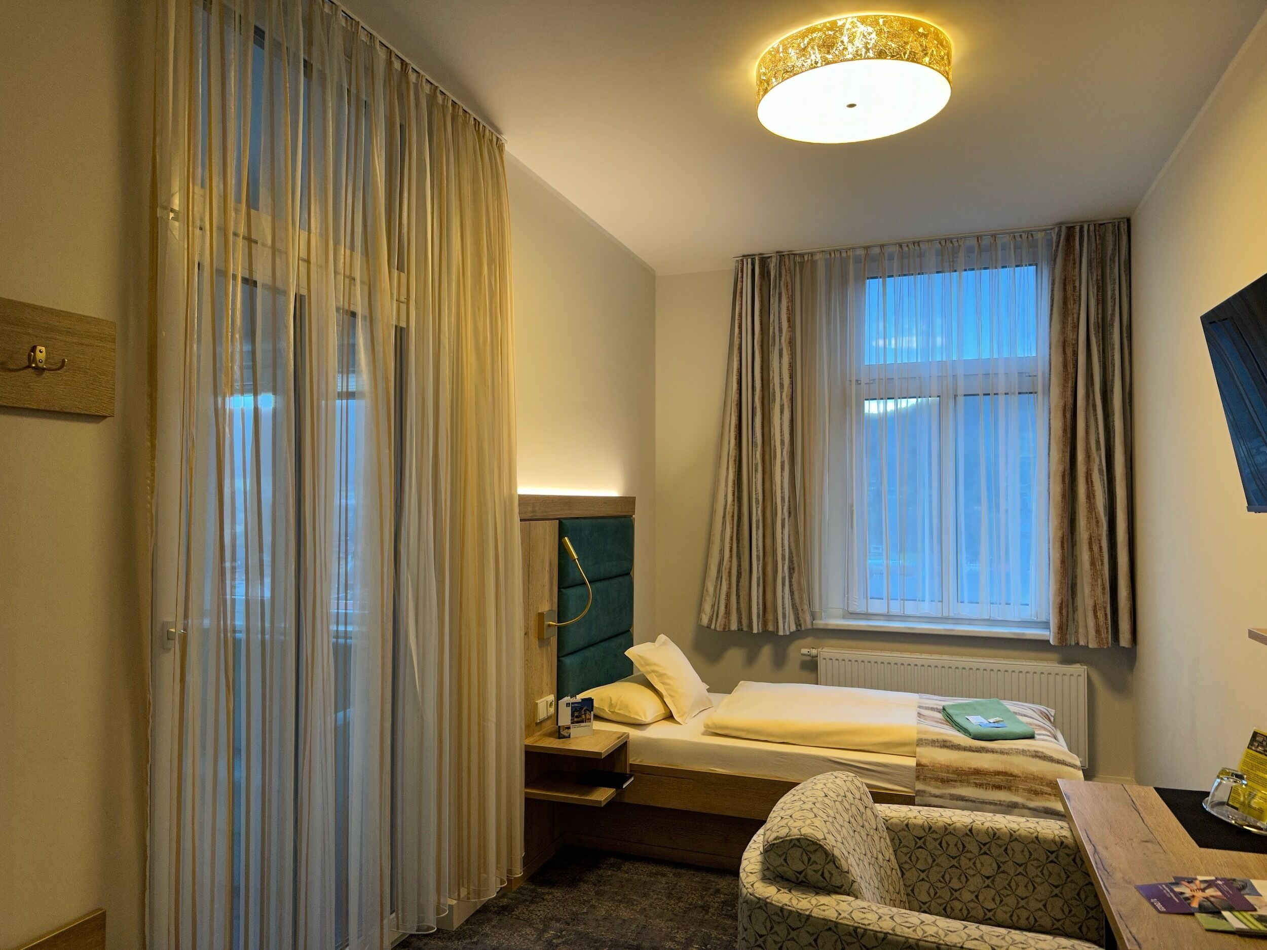 A hotel room with a bed, a desk, a chair, a window with curtains, a headboard, and a ceiling light.