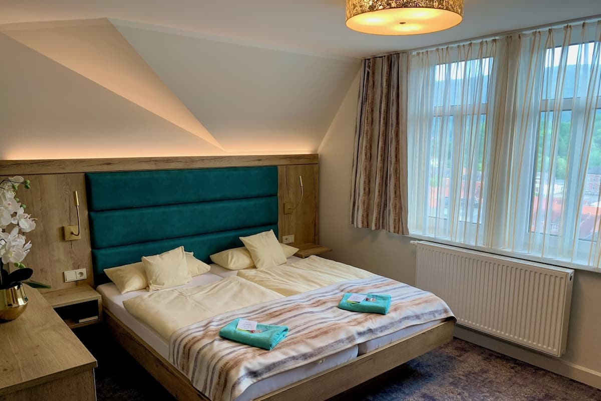 Un dormitorio con estructura de cama de madera, cabecera tapizada en color aguamarina y ventana con cortinas de gasa.