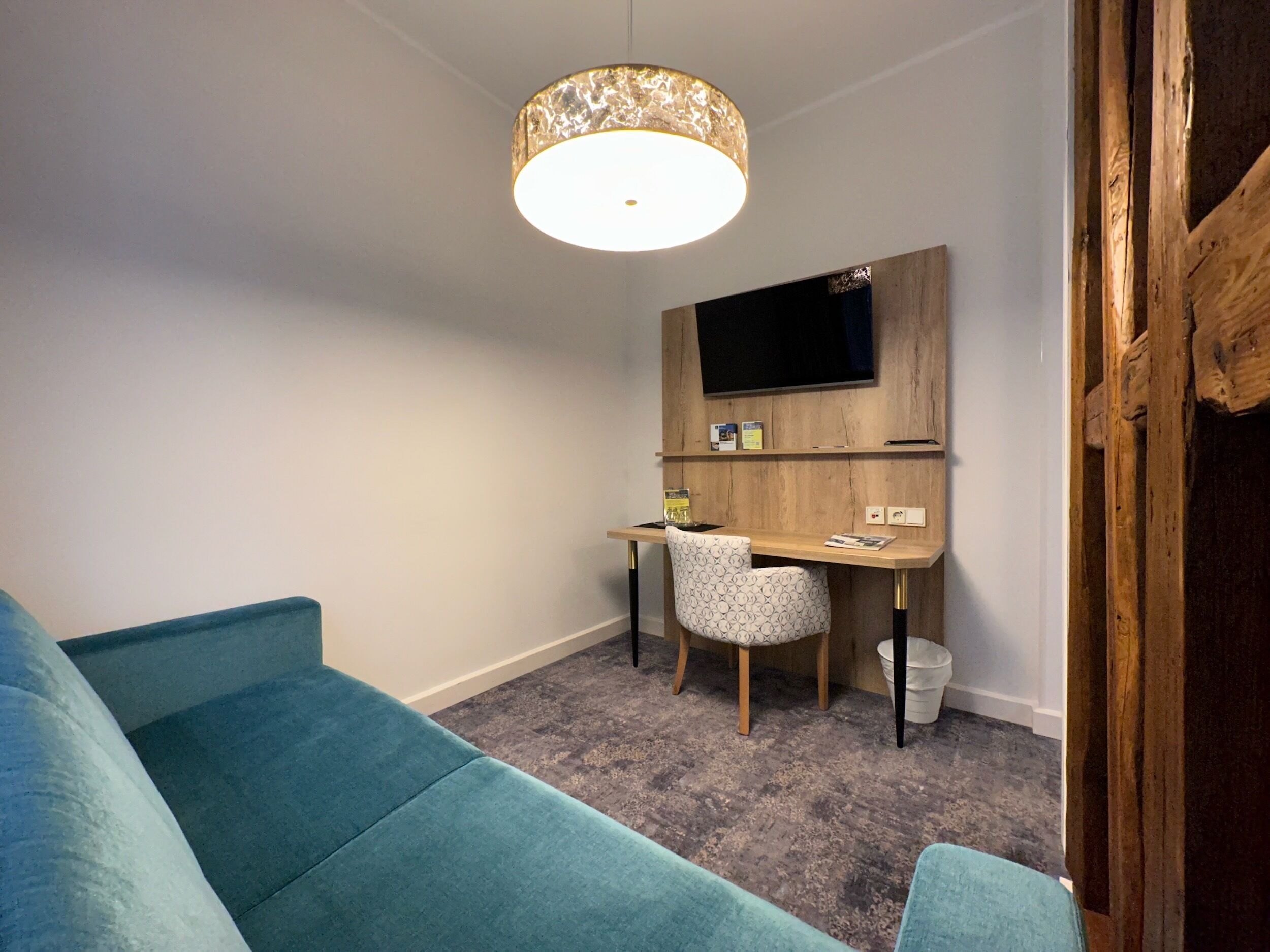 A compact living space with a teal sofa, a wooden desk with a chair, a mounted TV, and a patterned light fixture.