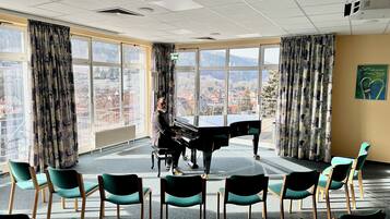Una habitación con un piano, sillas, ventanas amplias y vista a un paisaje urbano.