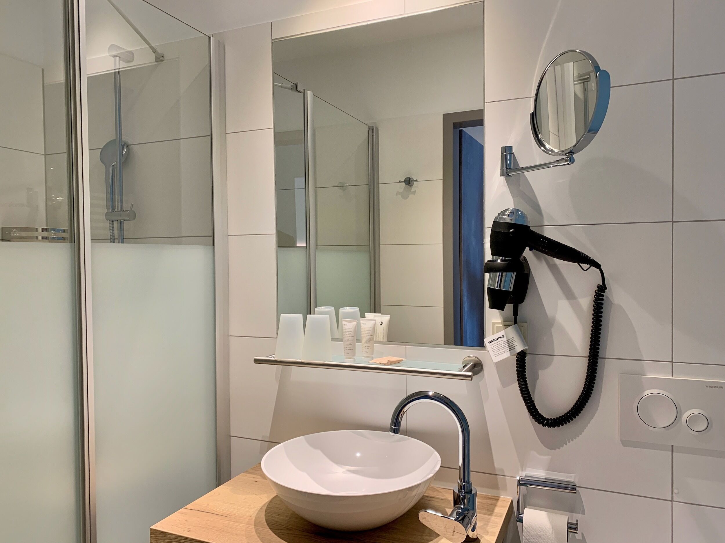 Ein modernes Badezimmer mit Duschkabine aus Glas, Waschbecken mit Holzablage, Spiegel und Wandhaartrockner.