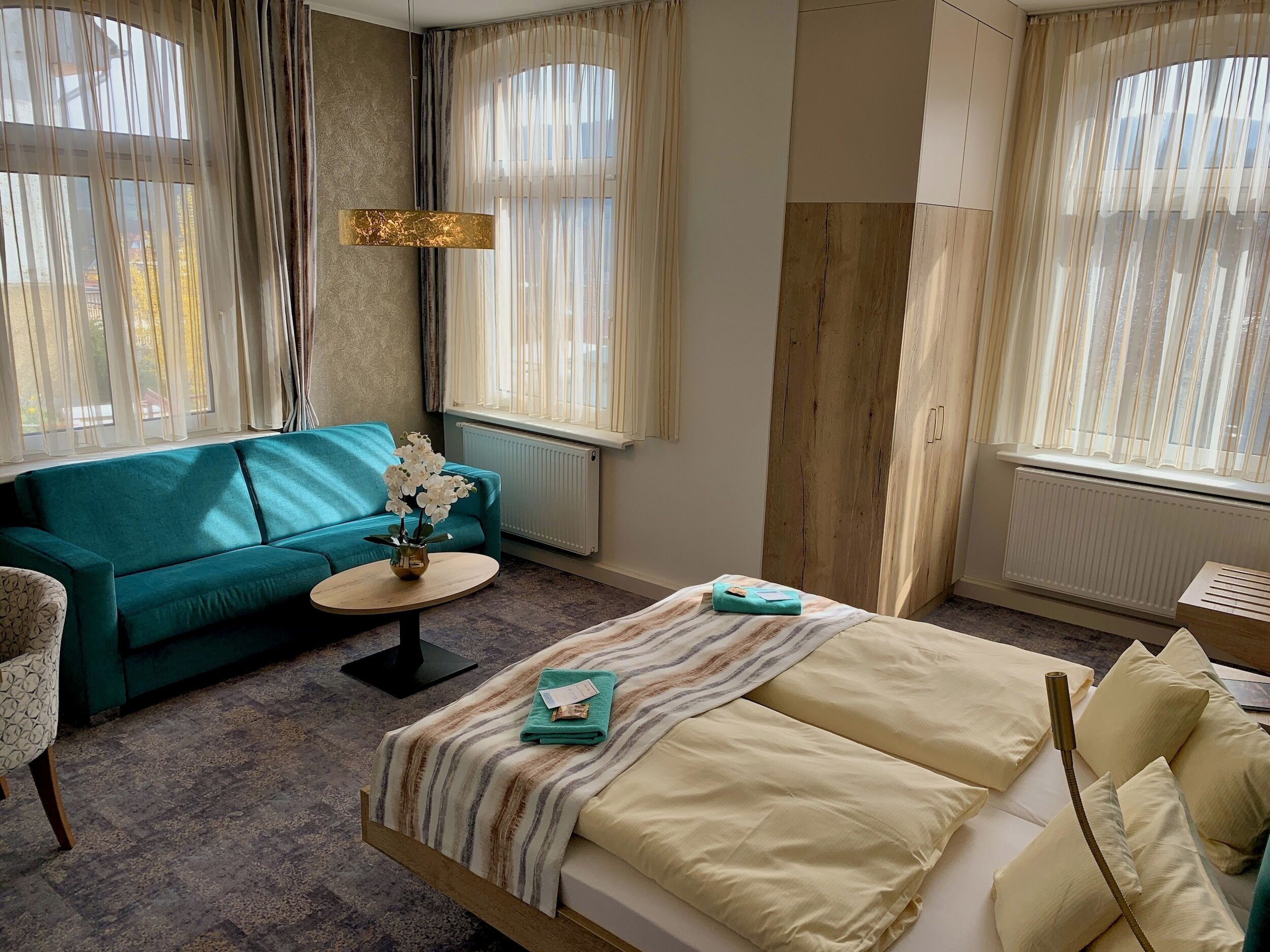 A hotel room with a bed, a sofa, a small table with a vase, and a window with curtains.